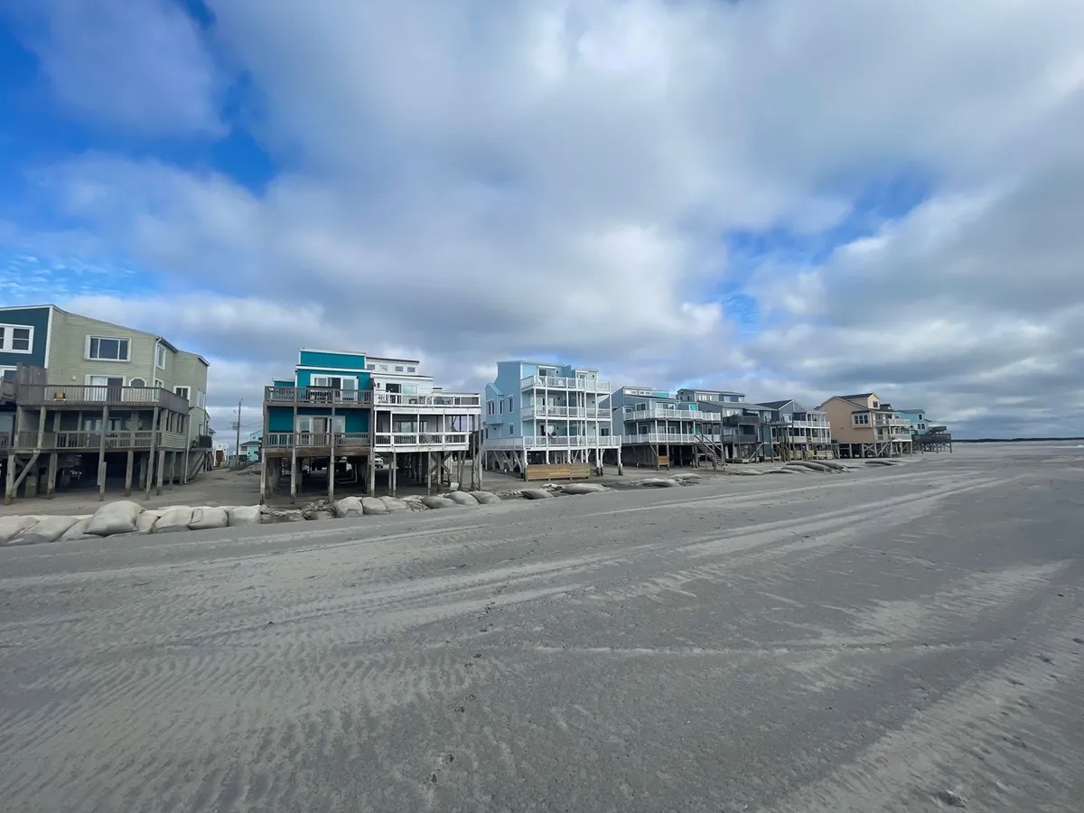 An exterior view showing colorful beach houses framed by sand dunes and a cloudy sky. The scene suggests a relaxed, communal environment, ideal for family gatherings and shared outdoor experiences.