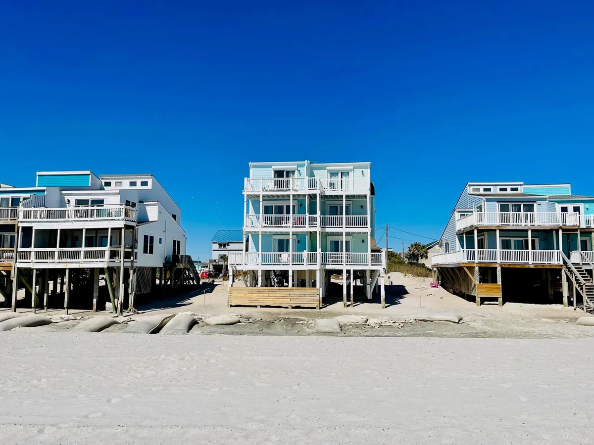 Exterior view of beachfront houses with spacious decks and balconies, a sand pathway leading directly to the beach. The scene radiates cheerfulness, and the decks provide ample space for families (and pets) to gather, sunbathe, or barbecue, with effortless transition from the home to the sand.