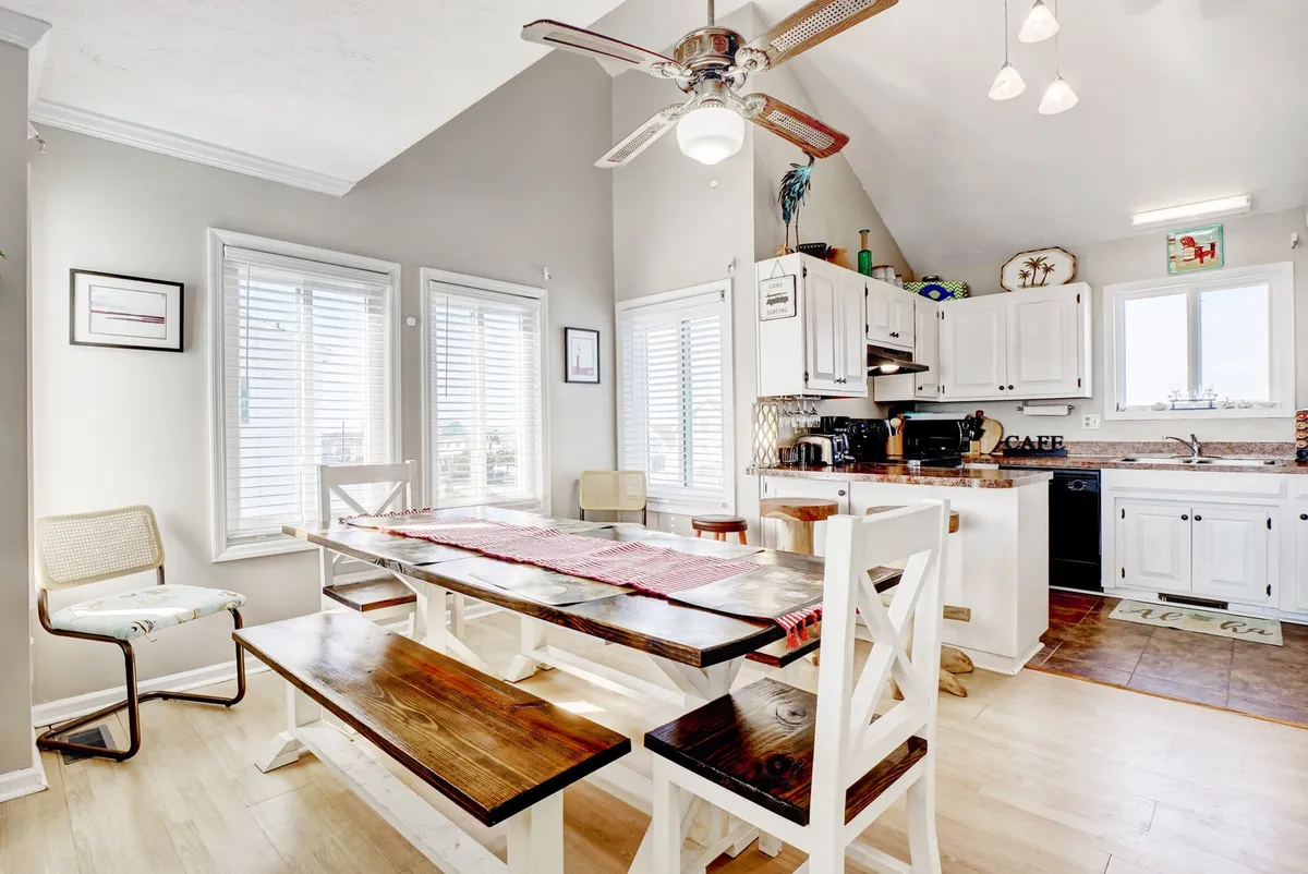 The dining area is centered around a large table flanked by window views, with the kitchen appliances and a ceiling fan visible. The scene suggests a welcoming setting for family meals, casual gatherings, and morning coffee, with warm, ambient lighting.