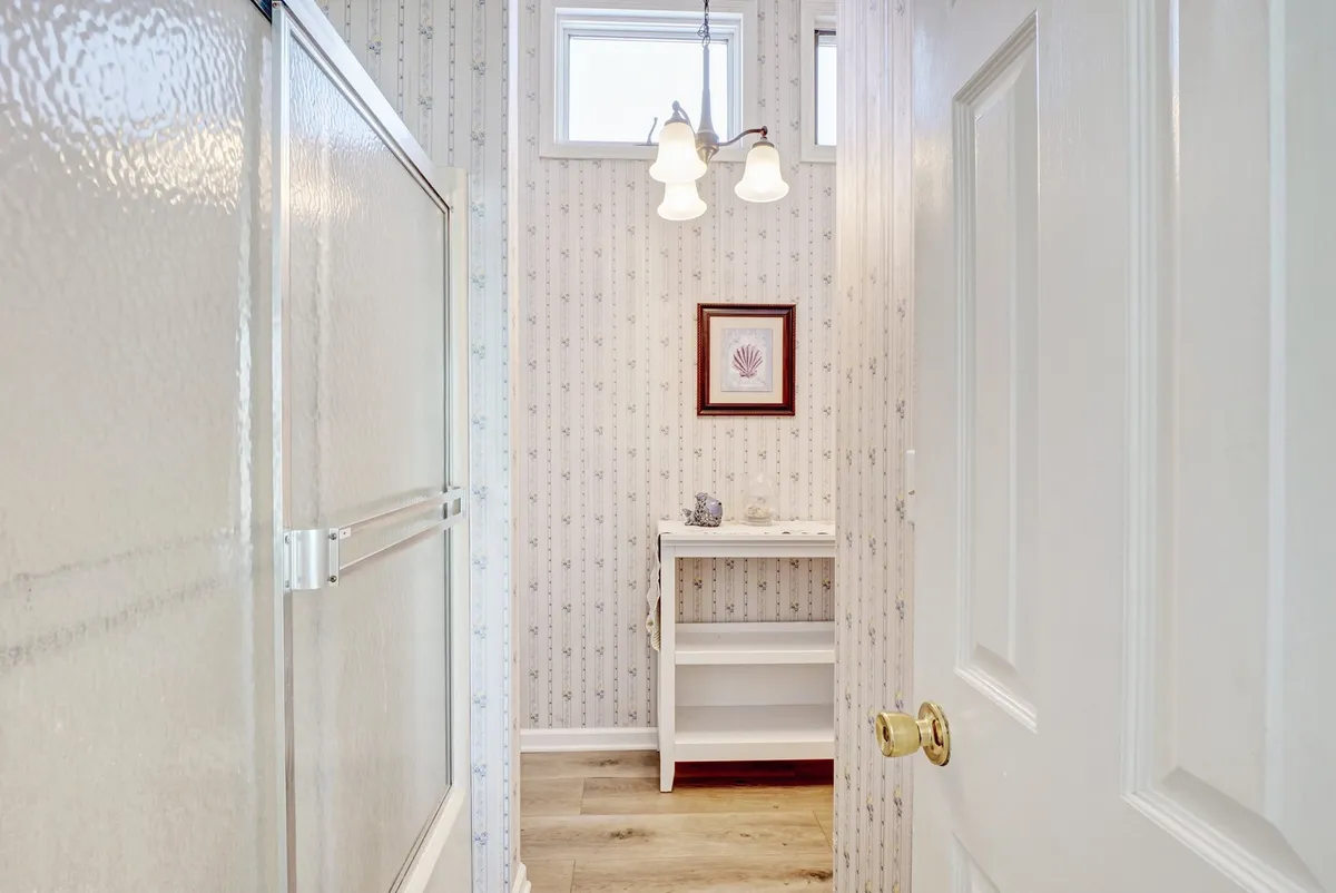 A calm, well-lit bathroom featuring a glass shower enclosure, framed shell artwork, and a vanity table. Gentle light streams in, creating a serene space ideal for relaxation and personal care.