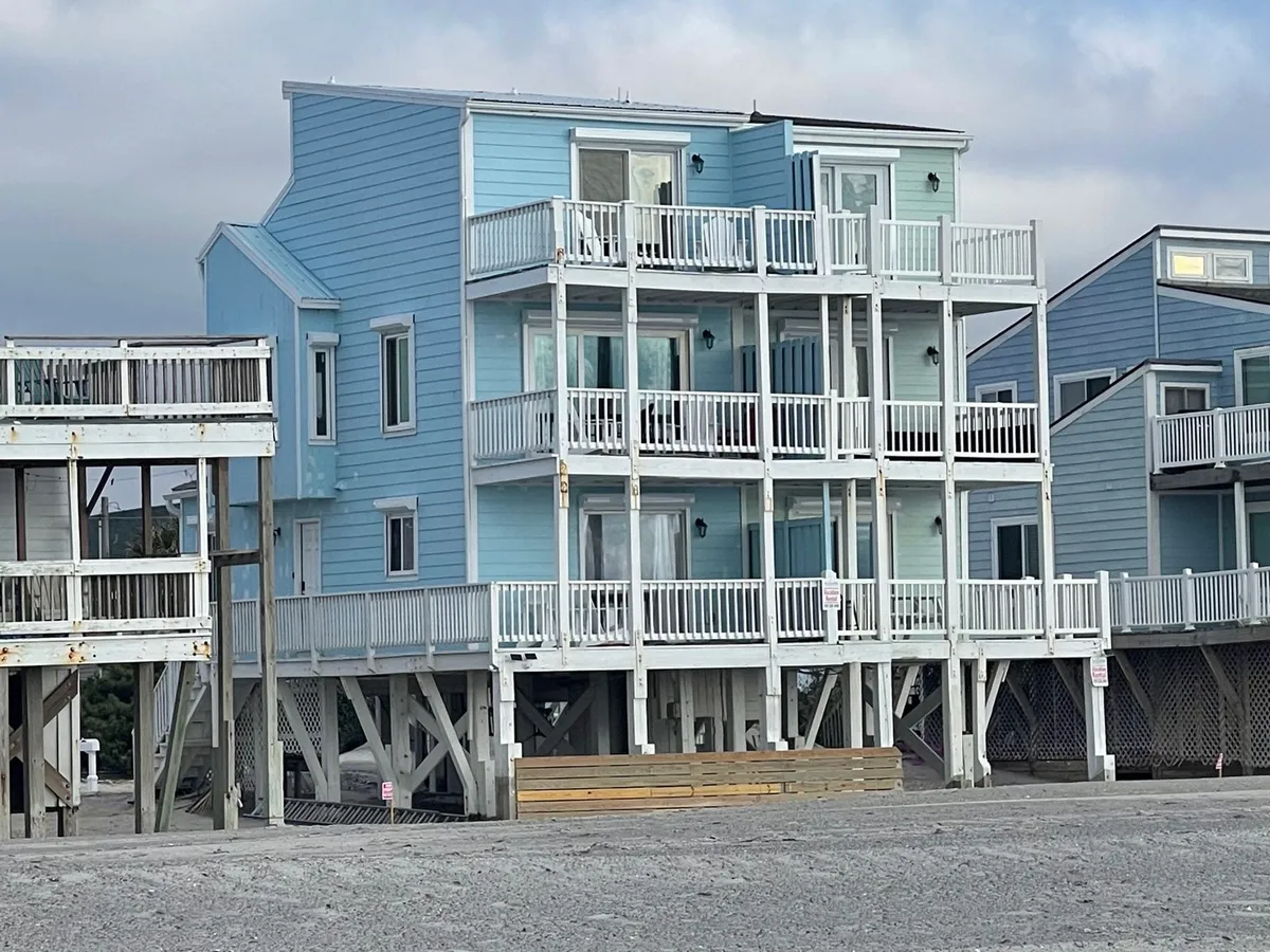 A striking blue beach house stands out with its bright white railings and multiple spacious balconies, offering plenty of outdoor space to relax and take in the coastal view. The balconies and decks provide a private retreat just steps from the sand, ideal for unwinding with pets in the fresh sea air.