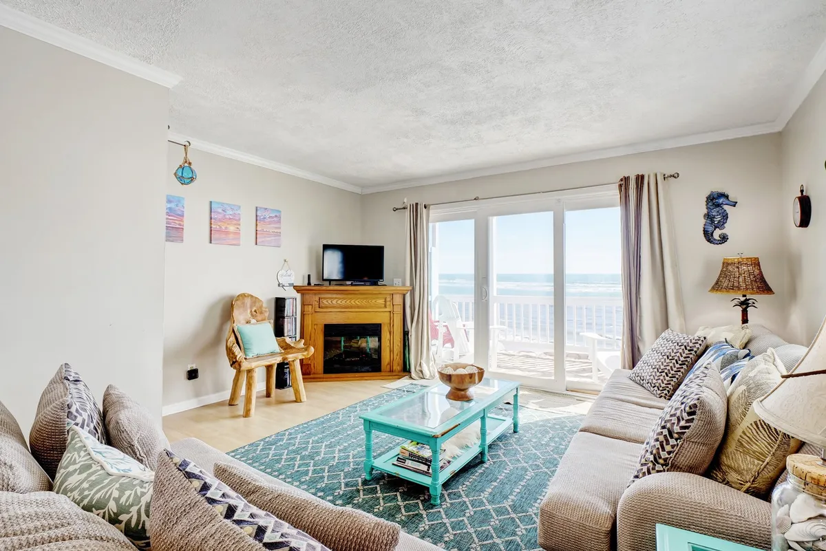 A cozy living area featuring large windows with an ocean view, teal coffee table, comfortable sofas, a decorative lamp, and colorful wall art. The layout encourages relaxation and togetherness, with abundant natural light and inviting decor.
