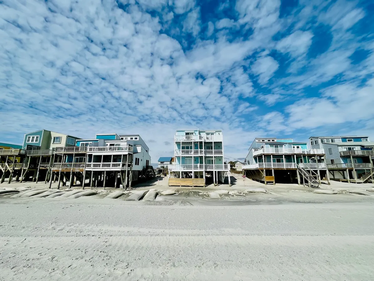 A wide exterior shot of several beach houses situated along a sandy shoreline under a cloudy sky. The image captures balconies facing the ocean, hints of the beach community atmosphere, and the inviting proximity of the beach itself.