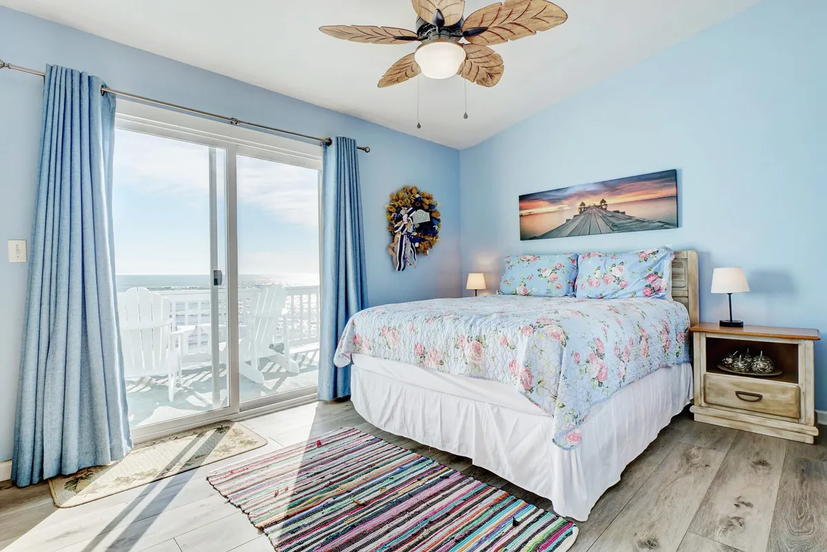 A bright, tranquil bedroom with floral bedding, a large window offering direct access to a balcony, a colorful area rug, and decorative wall art. The window frames expansive views, suggesting proximity to the ocean and creating a seamless connection between indoor comfort and the outdoor seascape.
