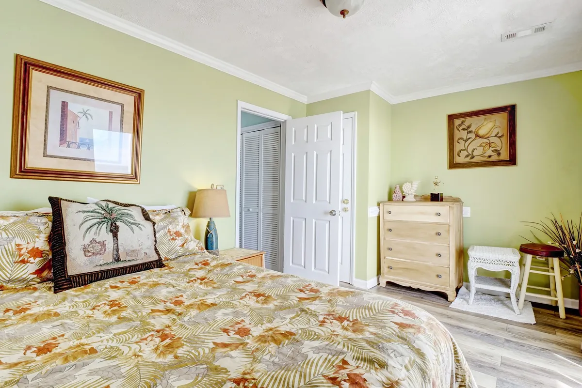 A serene bedroom featuring a bed with floral bedding, soft lamp lighting, framed artwork, and a welcoming chest of drawers. The setting is bright and tranquil, ideal for intimate moments and quiet relaxation.