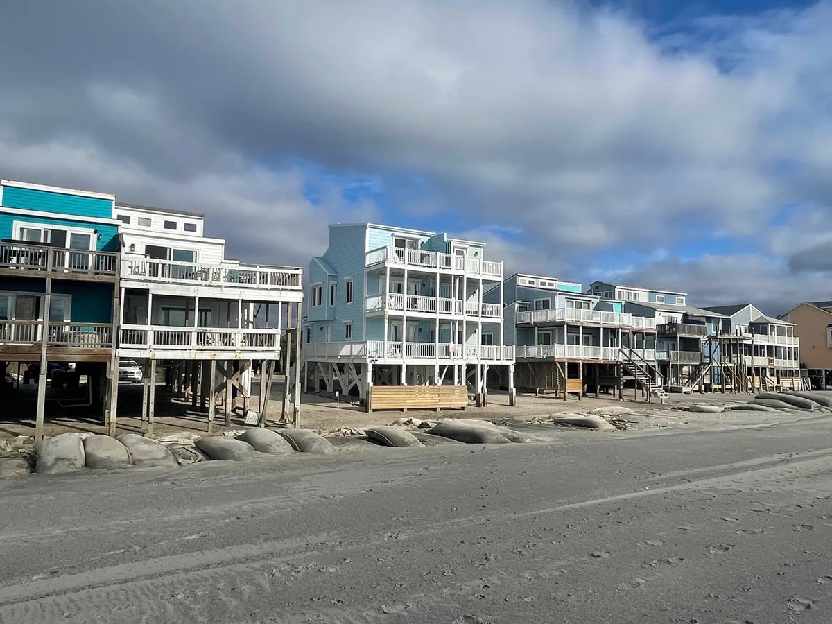 A charming row of beachfront houses, bordered by a sandy boardwalk and sand dunes, immersed in a tranquil coastal landscape. The inviting architecture and proximity to the shore suggest opportunities for beach walks and shared outdoor experiences.