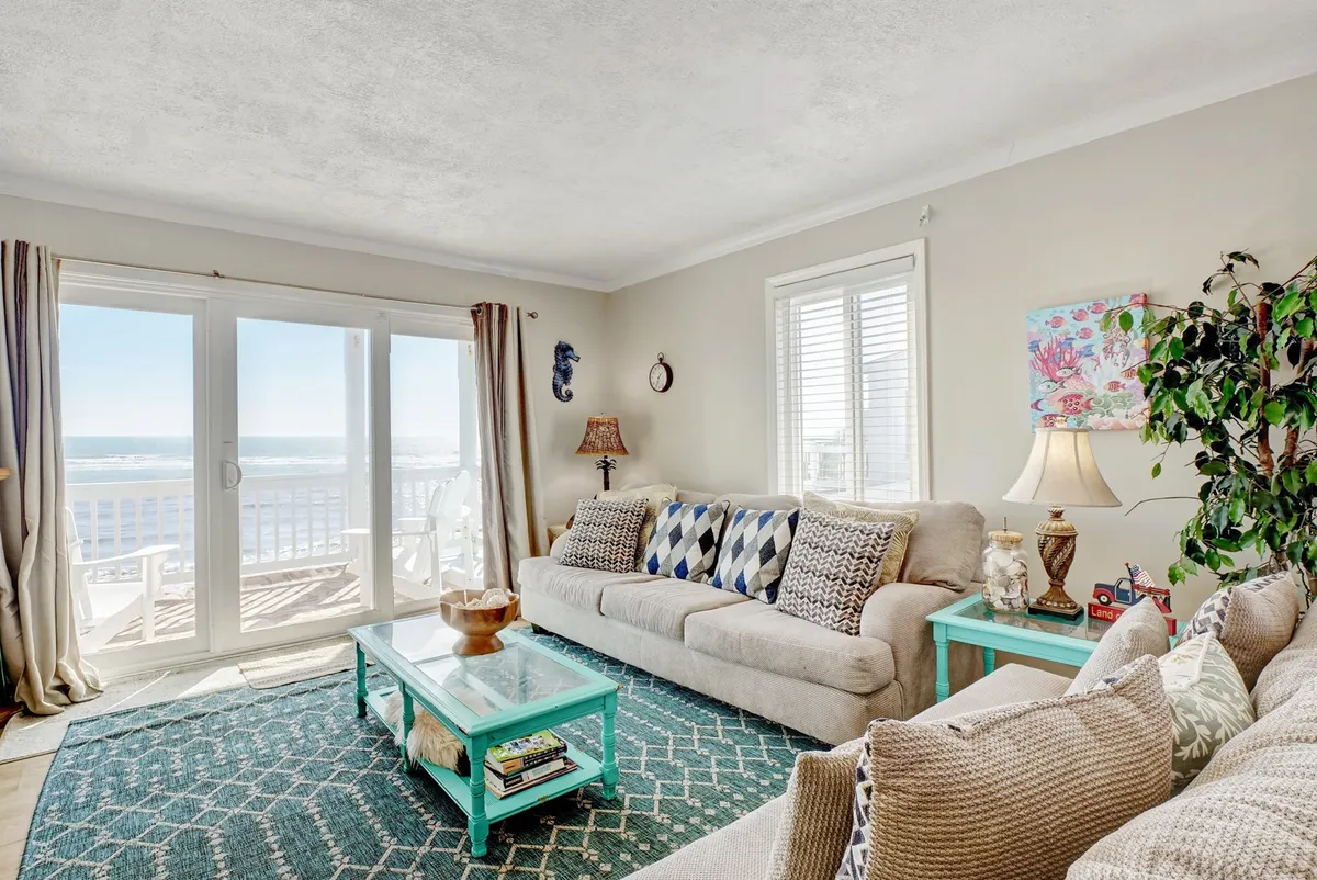 This interior living space features large windows with an ocean view, a comfortable sofa, a decorative coffee table, and colorful artwork. The abundance of natural light and the inviting arrangement of furniture create a welcoming spot to relax with family or pets, while still enjoying views of the beach.