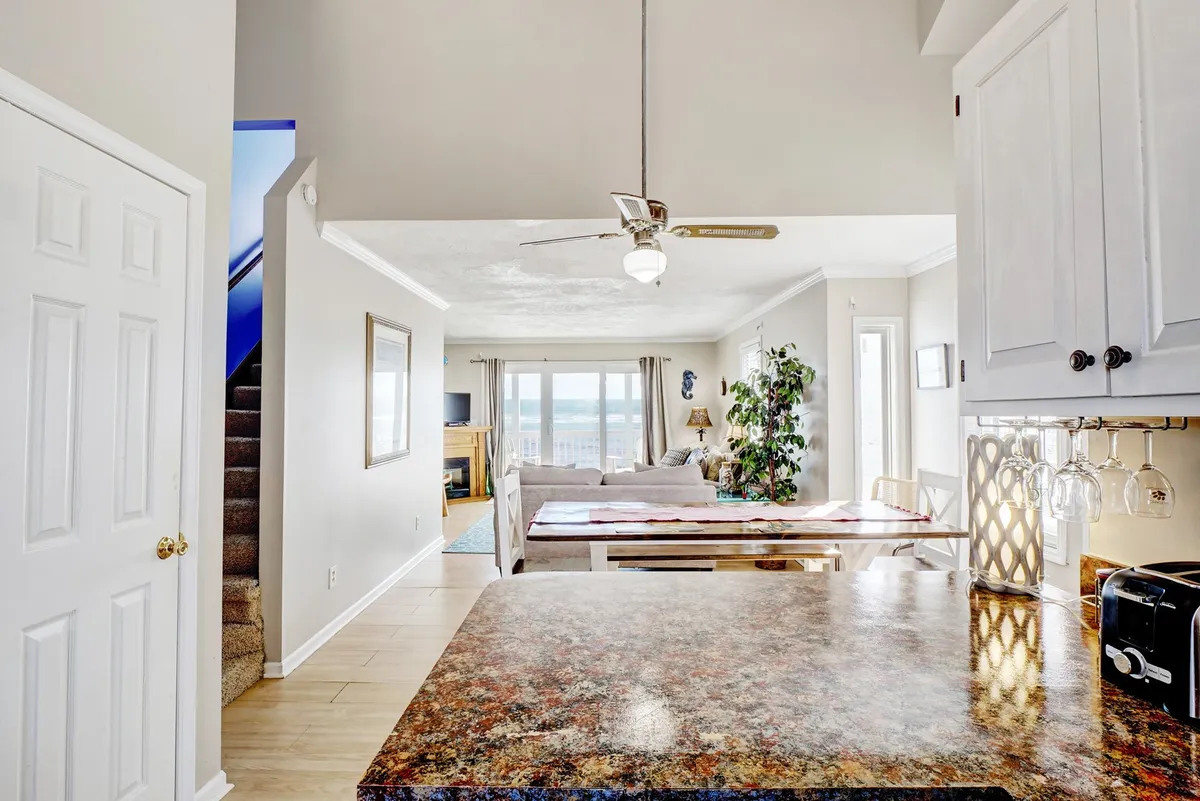 This image offers a wide-angle view of an open-plan area that seamlessly integrates the kitchen, dining table, living area with a sofa, and large windows framing a scenic view. The setup highlights the central role of this space as a communal hub, perfect for cooking, dining, and relaxing together while enjoying natural light and coastal vistas.