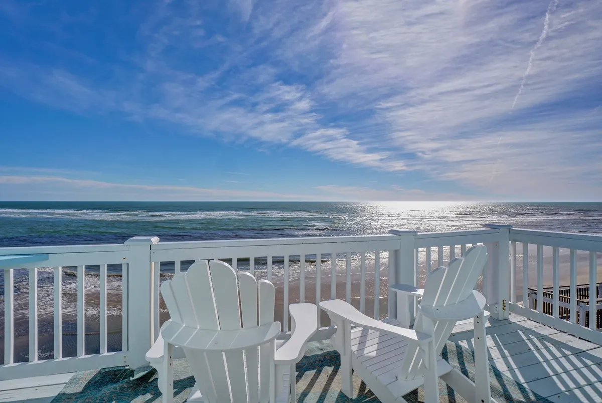 The image captures a sun-drenched deck overlooking the sparkling Atlantic, with inviting white Adirondack chairs perfectly positioned for panoramic ocean views. The gentle sunlight glimmers on the water, evoking a tranquil, coastal atmosphere ideal for unwinding while watching dolphins, reading a book, or soaking in the sunset. The setting exudes serene comfort and the promise of memorable gatherings on the shore.
