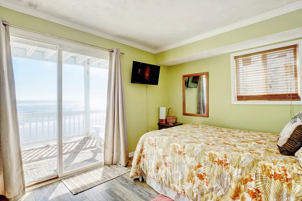 A bright queen bedroom featuring floral bedding, a large sliding glass door leading directly onto the deck, a mirror, and a wall-mounted TV. Sunlight pours into the room, and the inviting outdoor space is visible through the glass, creating a seamless transition between the bedroom and the deck.