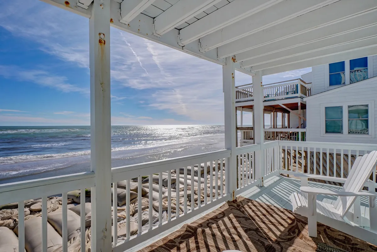 A tranquil oceanfront deck with white wooden railing and beach chairs, looking out over a panoramic ocean view. The setup is designed for relaxing, enjoying sunrises or sunsets, and soaking up the seaside atmosphere just outside the living spaces.